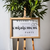 Invitation sign for Sam & Jonah's engagement party on a wooden stand with a plant in the background.